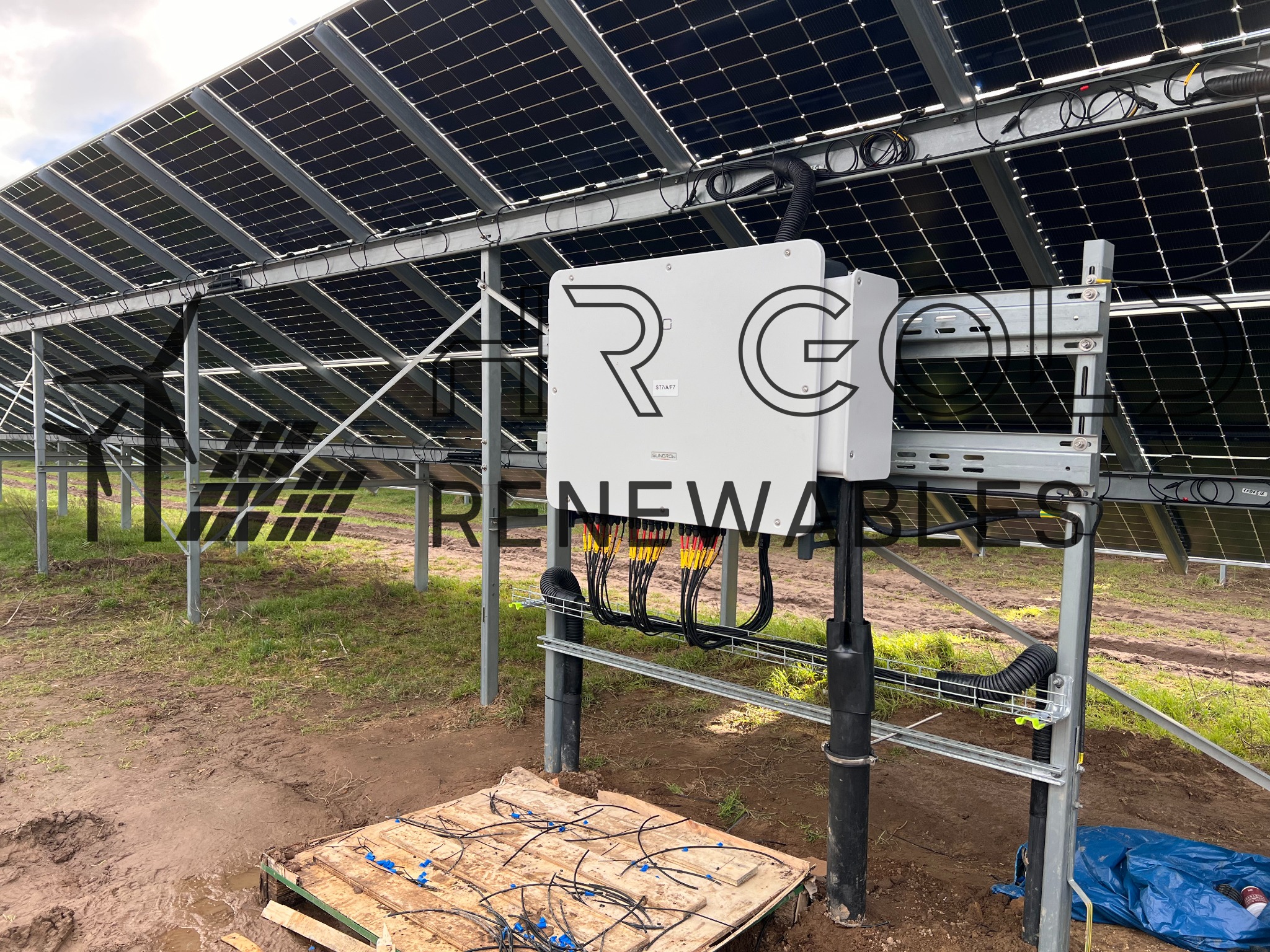 Instalacja paneli słonecznych: widok na inwerter z okablowaniem, na ziemi leżą kable i paleta, w tle rząd paneli słonecznych.