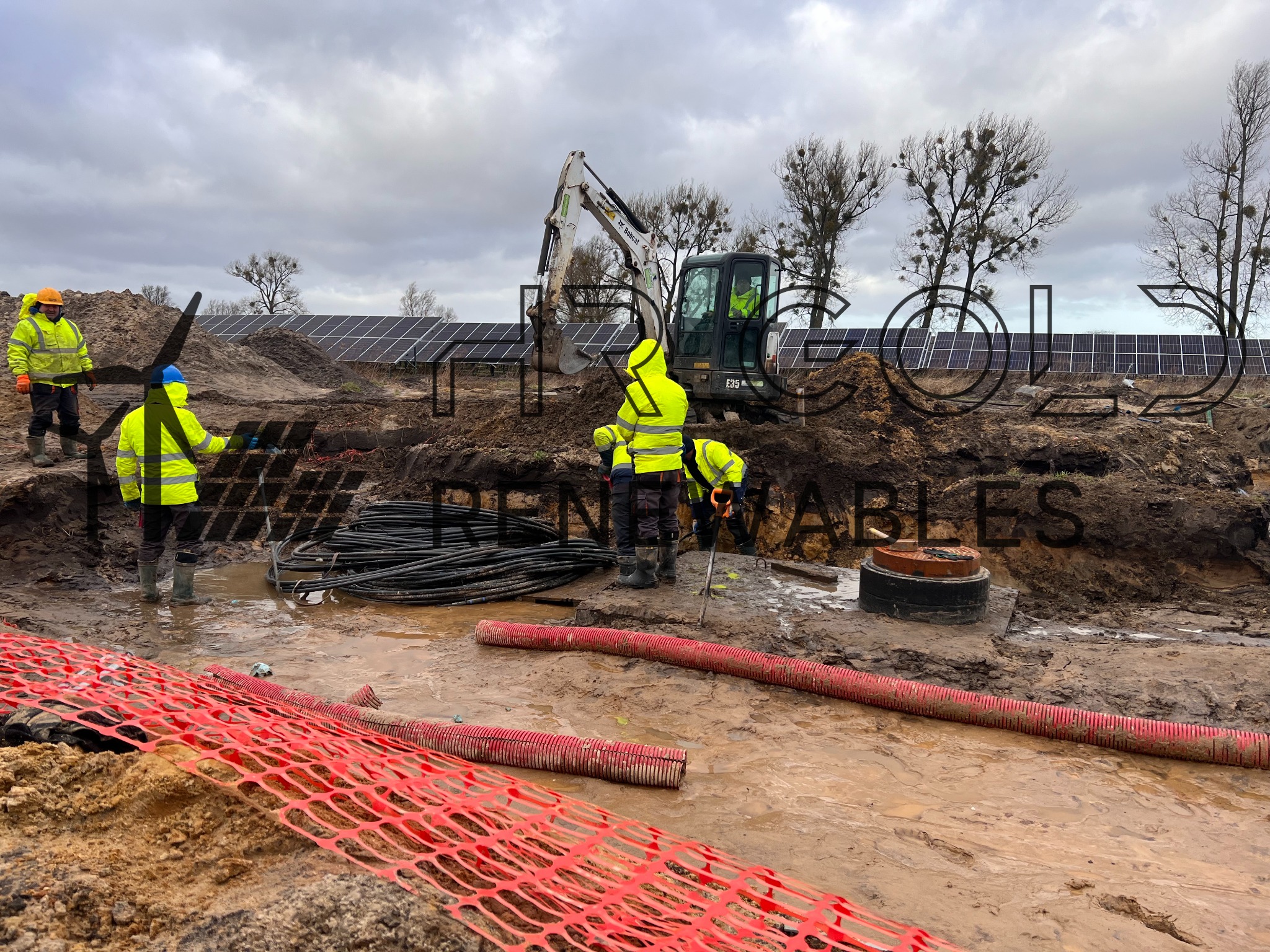 Instalacja paneli słonecznych: grupa pracowników w odblaskowych ubraniach pracuje przy wykopie, w tle widoczna koparka i rząd paneli fotowoltaicznych, na pierwszym planie czerwona siatka...