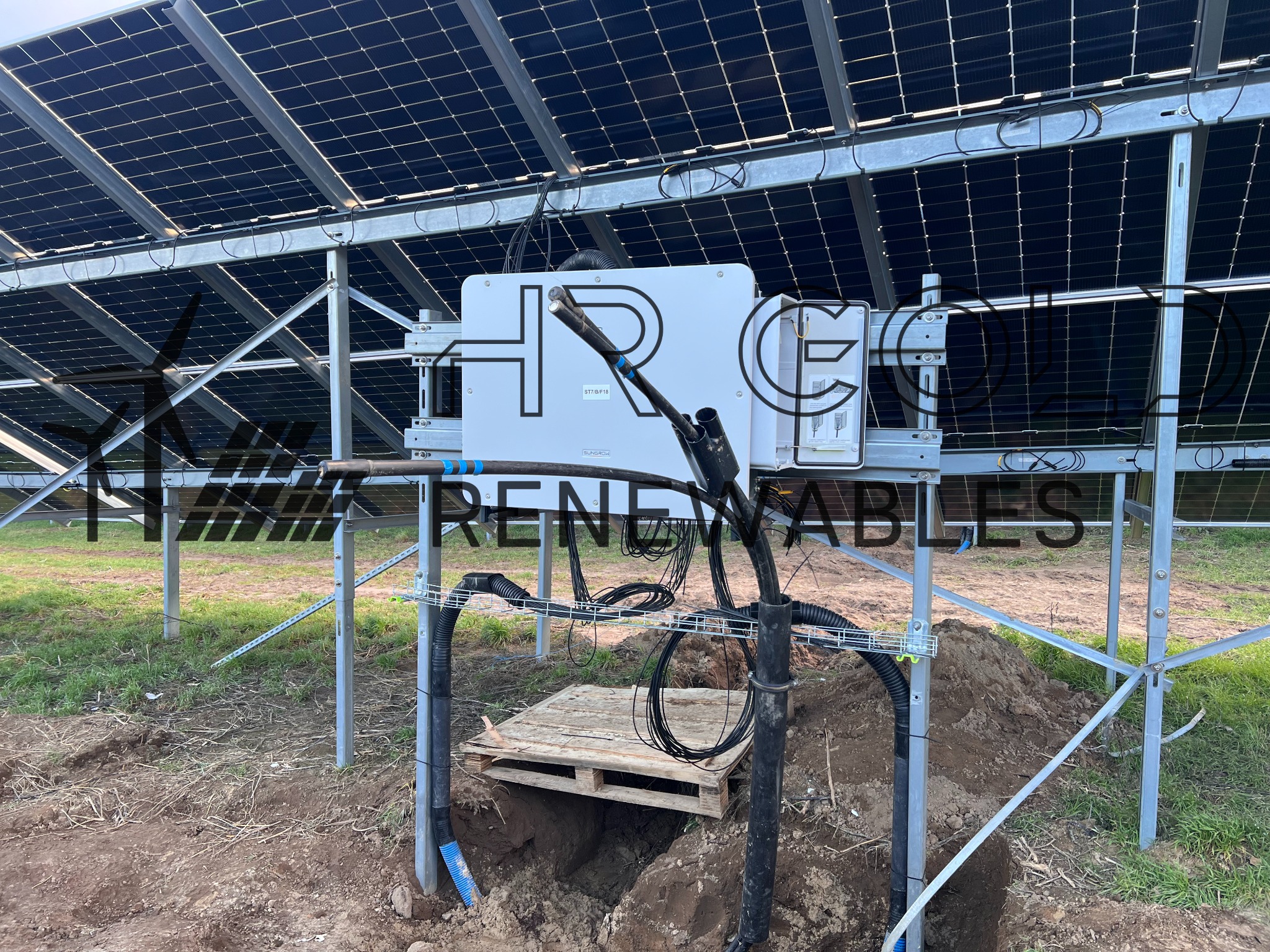 Instalacja paneli fotowoltaicznych w trakcie montażu: widoczne panele na metalowej konstrukcji, skrzynka elektryczna, kable prowadzone w ziemi i drewniana paleta zakrywająca wykop.