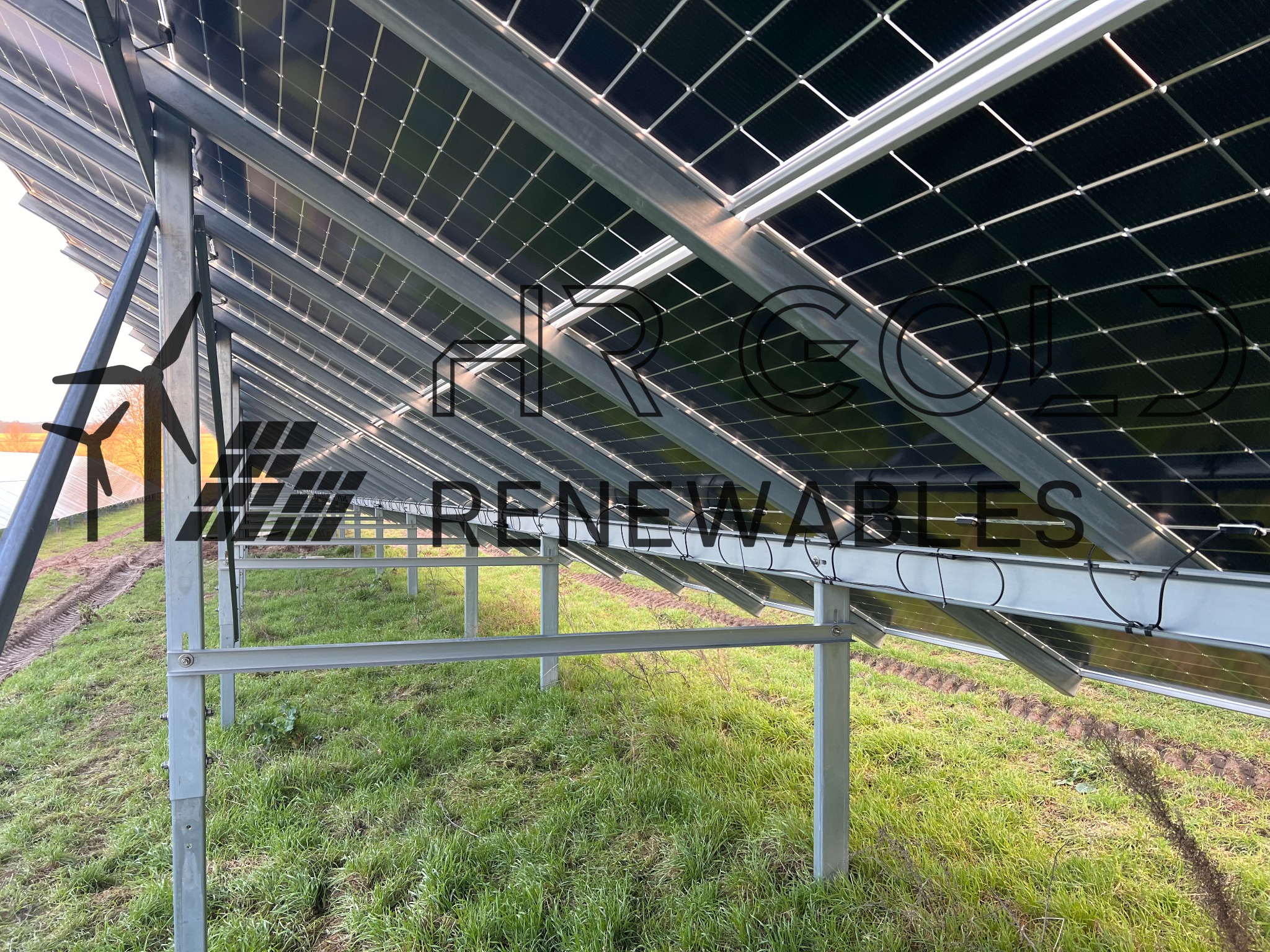 Instalacja paneli fotowoltaicznych na metalowej konstrukcji, widok od spodu, trawa i ślady opon w tle.