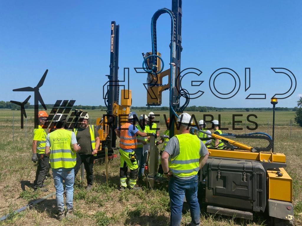 Grupa pracowników w kamizelkach odblaskowych obsługuje specjalistyczną maszynę do wbijania pali w terenie otwartym, prawdopodobnie pod instalację farmy wiatrowej.