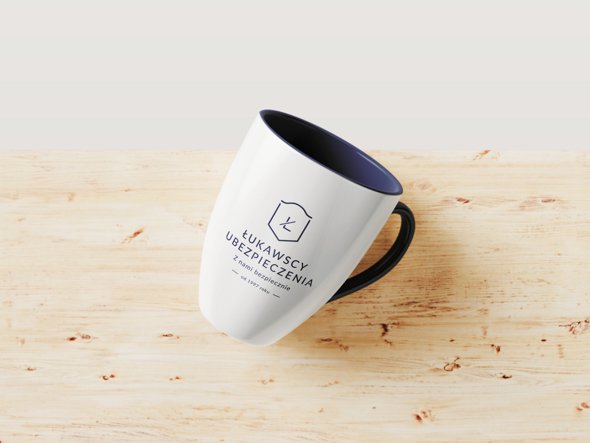 Biały kubek z granatowym wnętrzem i czarnym uchem, leżący ukośnie na jasnym drewnianym blacie. Na kubku widnieje czarny logotyp i napis 'ŁUKAWCY UBEZPIECZENIA. Z nami bezpiecznie - od 1997 roku'.