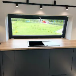 Nowoczesna kuchnia z czarnymi szafkami i drewnianym blatem, okno nad zlewem z widokiem na zielony ogród, ekspres do kawy na blacie.