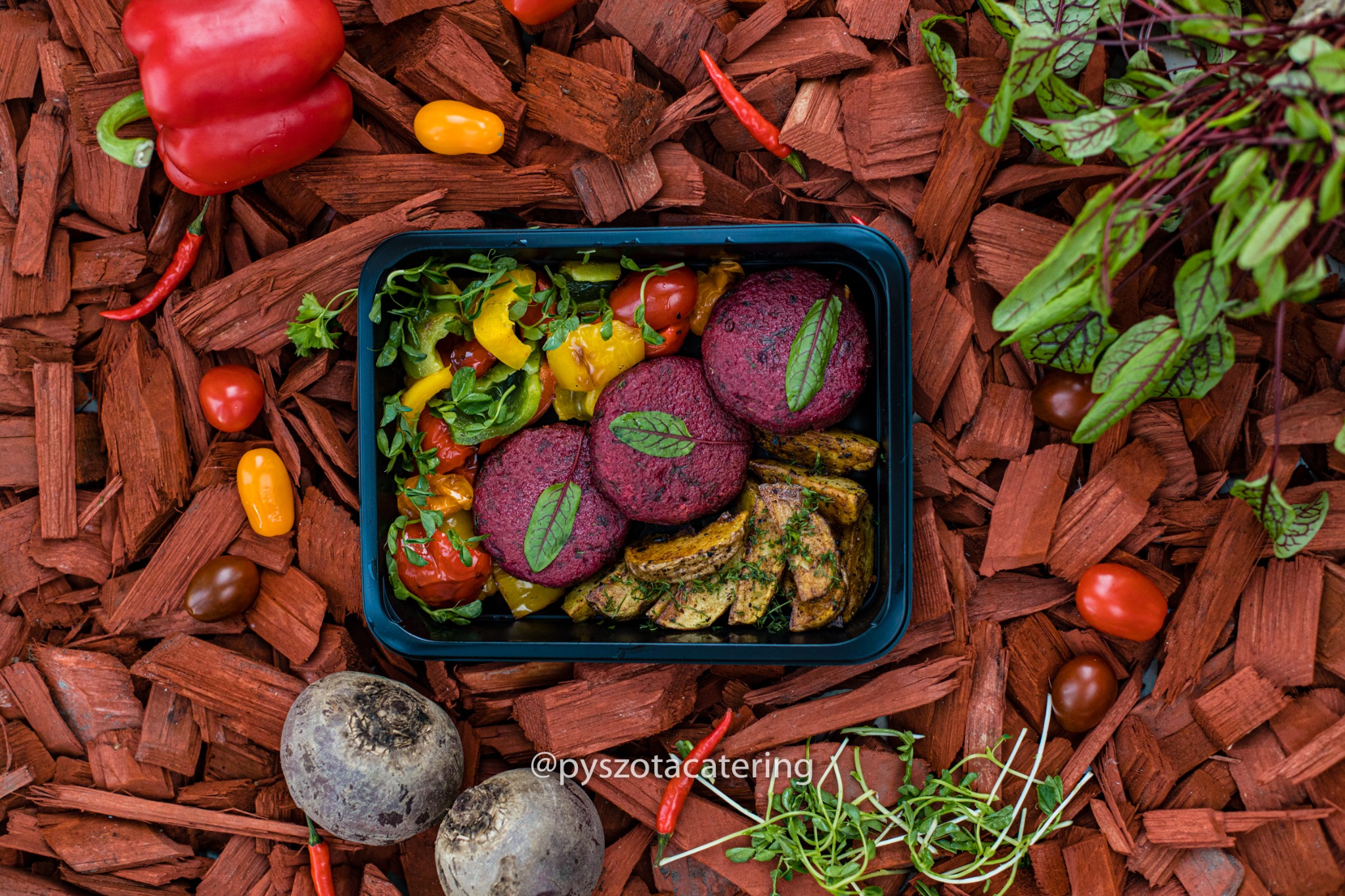 Gotowe danie dietetyczne w czarnym pojemniku na tle kory drzewnej: kotlety z buraków z liściem, pieczone ziemniaki z ziołami, grillowana papryka i pomidorki koktajlowe. Całość dopełniają świeże...