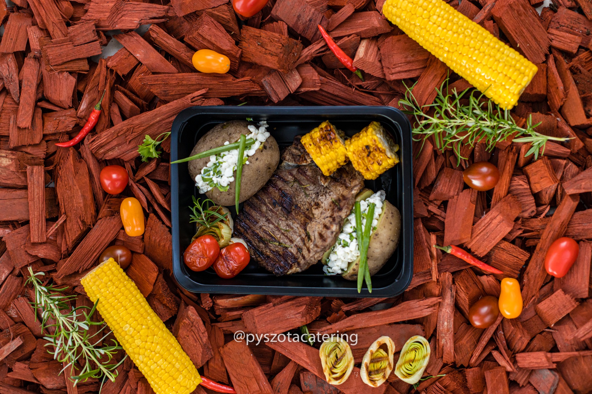 Cateringowe danie w czarnym pojemniku: grillowany stek, pieczone ziemniaki z twarożkiem i szczypiorkiem, grillowana kukurydza, pomidorki koktajlowe, całość na tle z czerwonych wiórów drzewnych,...