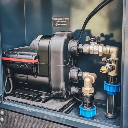 PROWIERT SPÓŁKA Z OGRANICZONĄ ODPOWIEDZIALNOŚCIĄ - Czarna pompa Grundfos w szarej obudowie z rurami i zaworami, instalacja do wiercenia studni. Widoczna tabliczka informacyjna firmy Pro-Wiert-Bud.