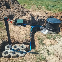 PROWIERT SPÓŁKA Z OGRANICZONĄ ODPOWIEDZIALNOŚCIĄ - Instalacja studni głębinowej: czarna skrzynka, niebieska rura, kran z czerwoną rączką, obudowa z pokrywą, kostka brukowa, wykop w ziemi, trawa.