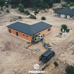 PROWIERT SPÓŁKA Z OGRANICZONĄ ODPOWIEDZIALNOŚCIĄ - Wiercenie studni głębinowej przy nowym domu jednorodzinnym. Widok z drona na plac budowy, gdzie ekipa montuje sprzęt wiertniczy obok budynku w stanie surowym.