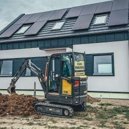 PROWIERT SPÓŁKA Z OGRANICZONĄ ODPOWIEDZIALNOŚCIĄ - Kompaktowa koparka Volvo EC18E w trakcie prac ziemnych na działce z nowoczesnym domem z panelami słonecznymi na dachu. Widoczny operator w kabinie.