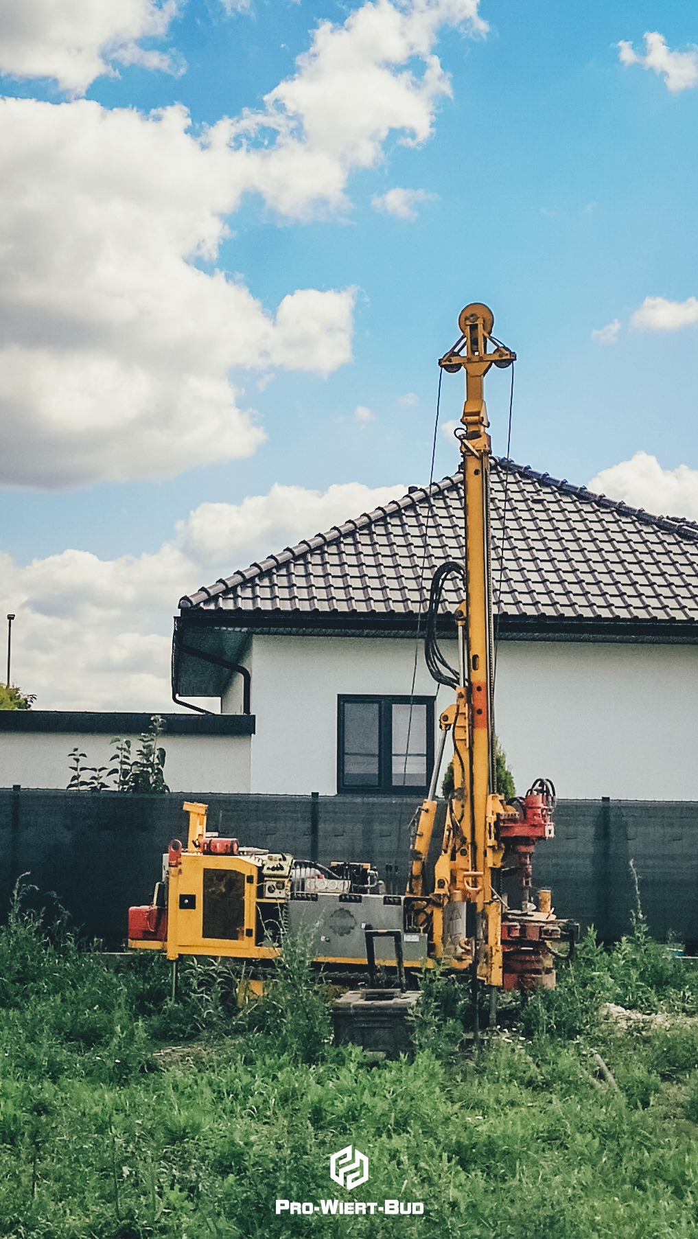Żółta wiertnica na tle domu z ciemną dachówką i błękitnego nieba z chmurami, otoczona zieloną trawą i szarym ogrodzeniem.