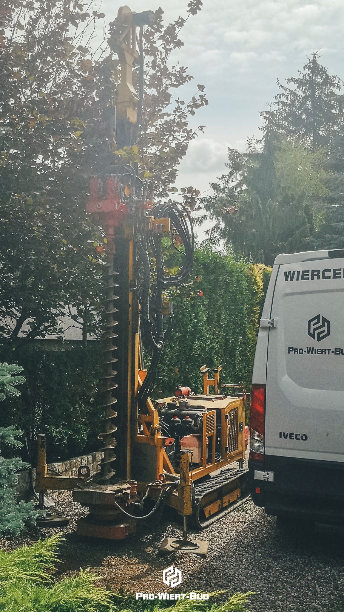 Żółta wiertnica na gąsienicach, częściowo zasłonięta przez drzewo, obok biały samochód dostawczy z nazwą firmy wiertniczej.