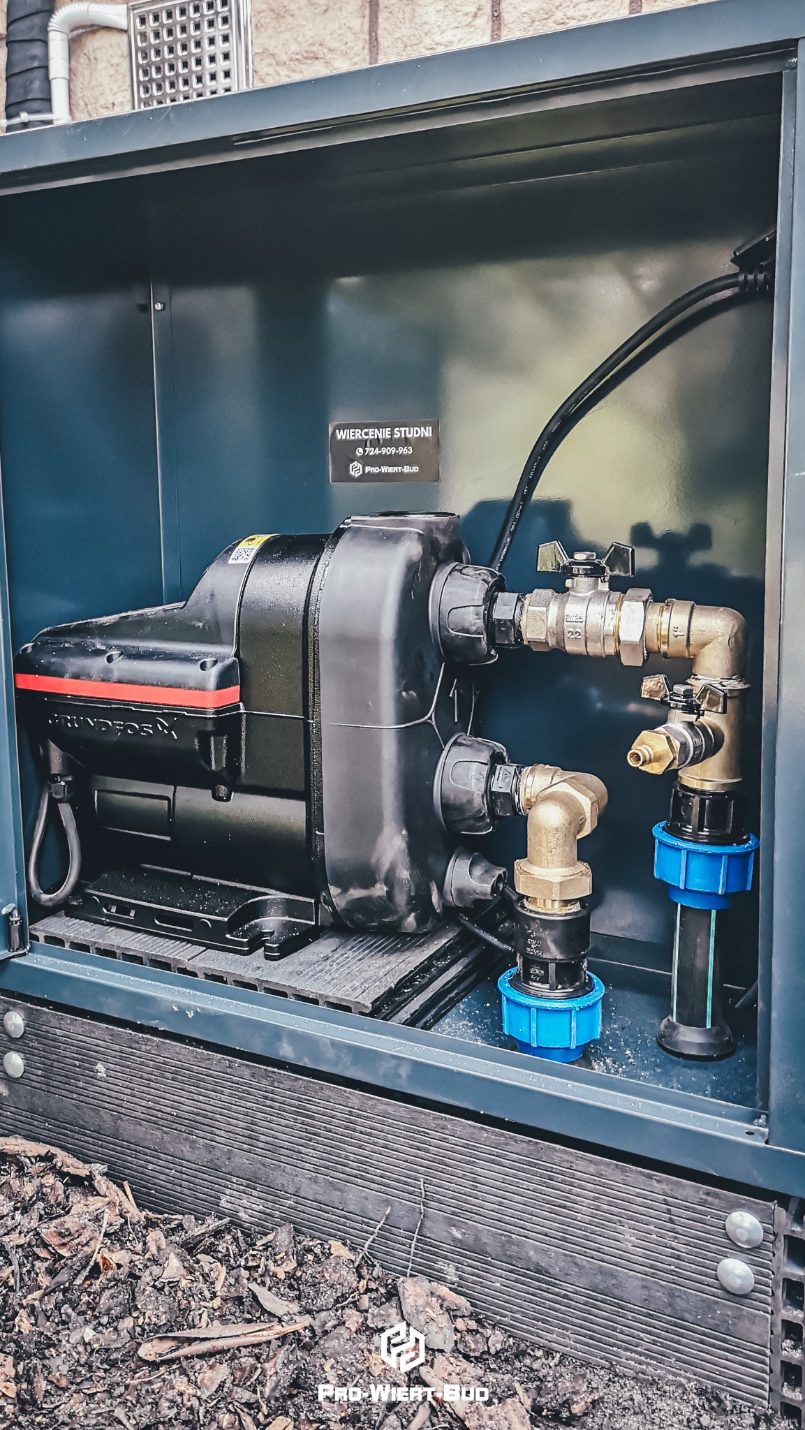 Czarna pompa Grundfos wewnątrz szarej obudowy, z widocznymi przyłączami i zaworami, na tle drewnianego podłoża w otoczeniu ściółki.