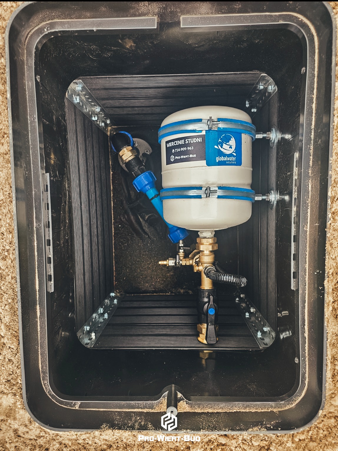 Zabezpieczona studnia głębinowa w czarnej skrzynce z widocznym zbiornikiem hydroforowym Global Water Solutions oraz elementami instalacji wodnej.