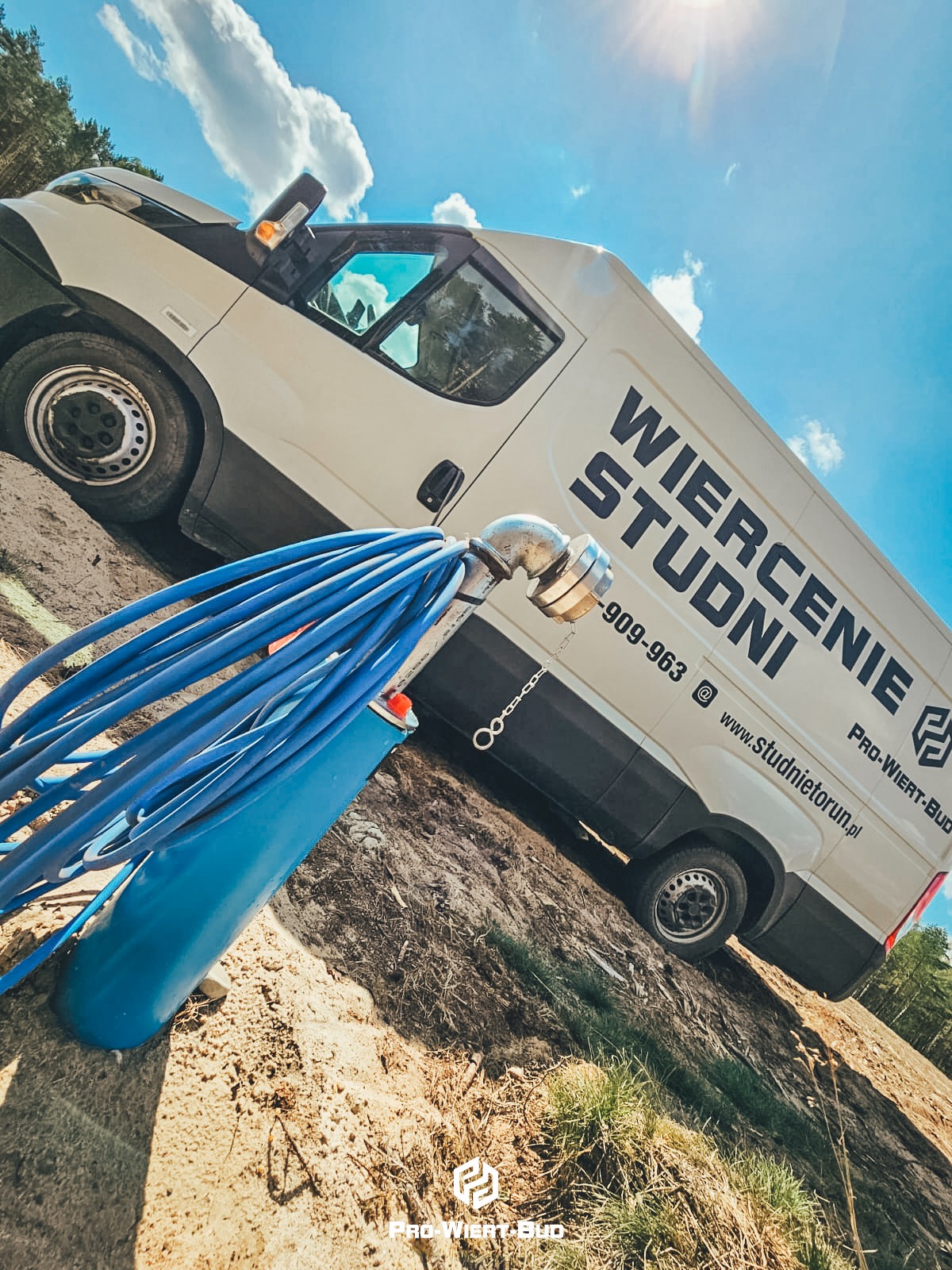 Węże do pompowania wody z nowo wywierconej studni w Toruniu, na tle samochodu firmy wiertniczej z widocznym logo i numerem telefonu.