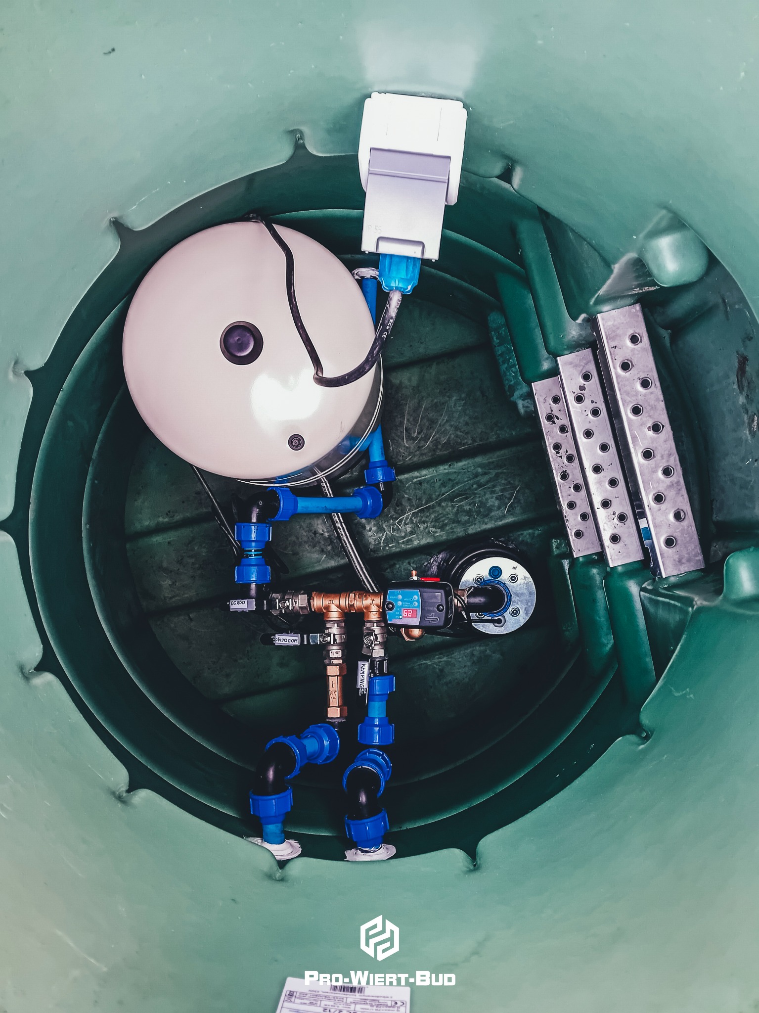 Widok z góry na instalację hydroforową w zielonym kręgu studni, z białym zbiornikiem ciśnieniowym, niebieskimi rurami, manometrem i metalowymi elementami mocującymi.