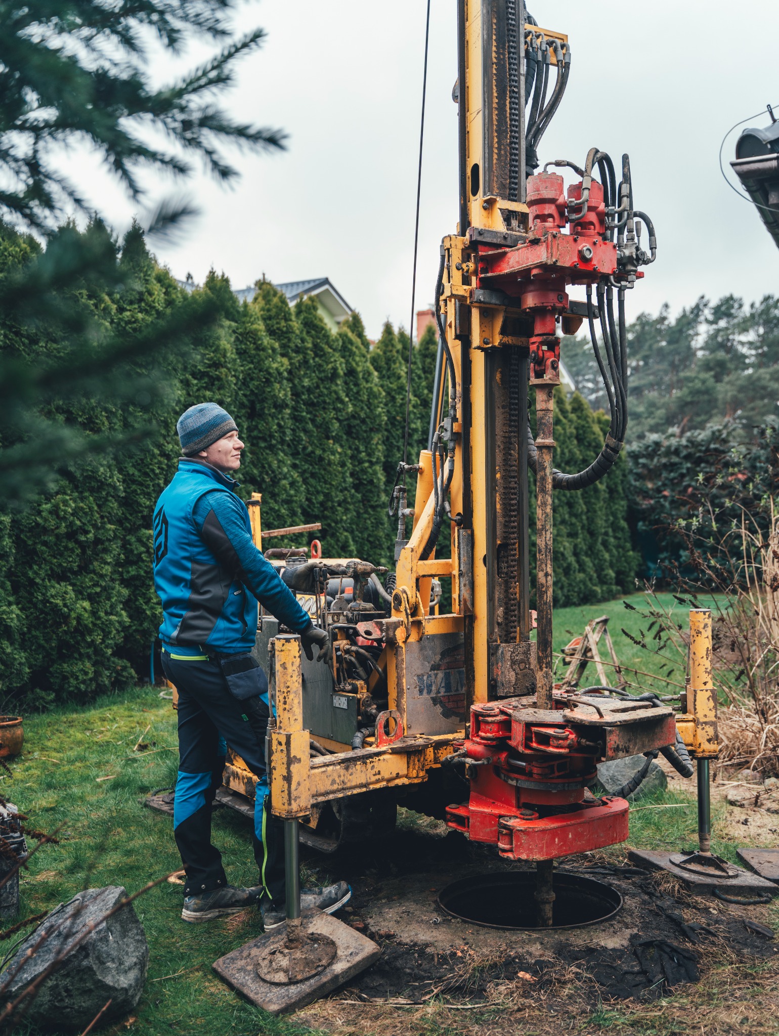 Pracownik w niebieskiej kurtce obsługuje żółto-czerwoną wiertnicę na trawniku, wiercenie otworu w ziemi, w tle iglaki i dom.
