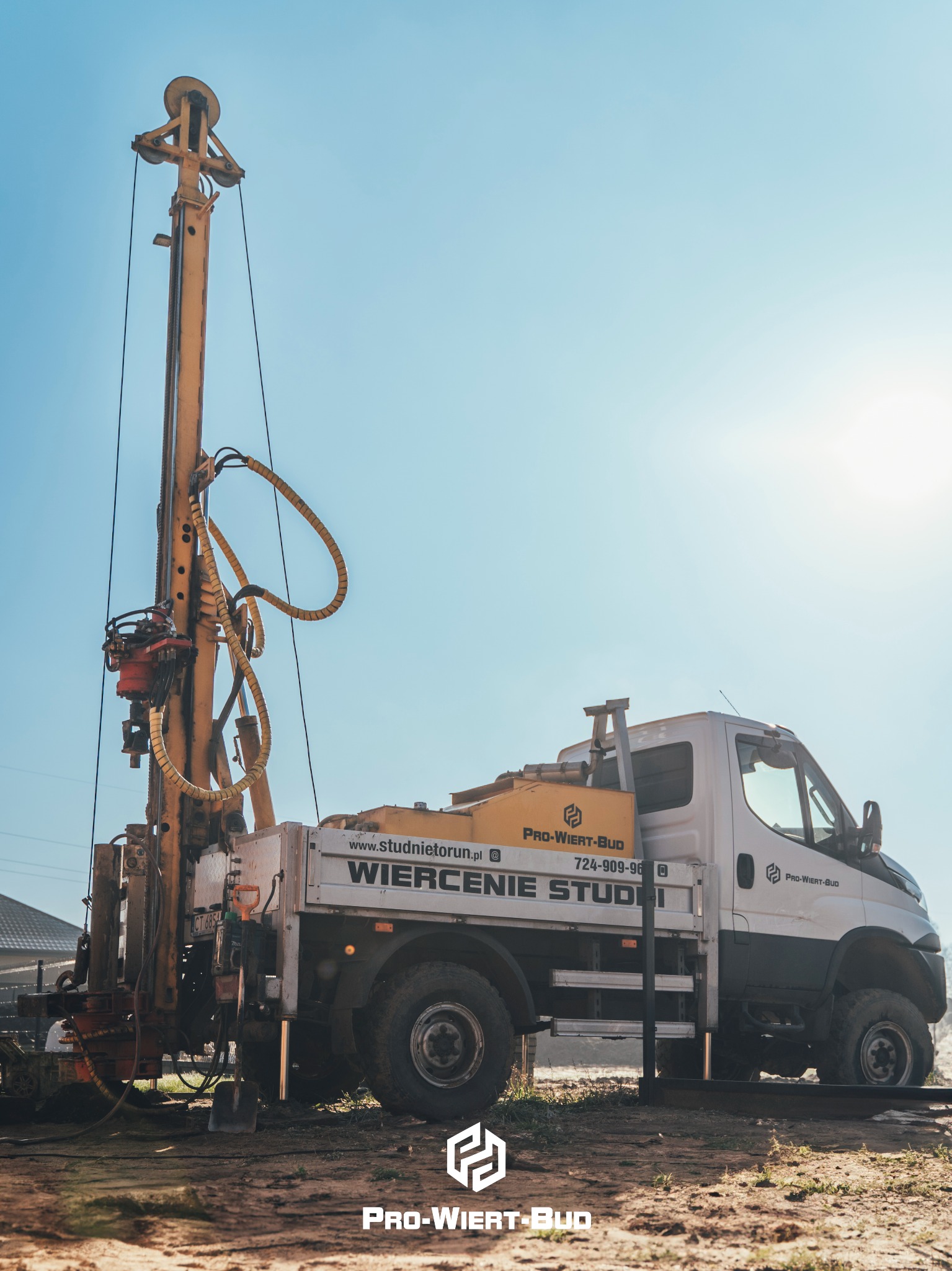 Mobilna wiertnica studni na podwoziu samochodu ciężarowego, z widocznym osprzętem wiertniczym i elementami hydraulicznymi na tle błękitnego nieba.