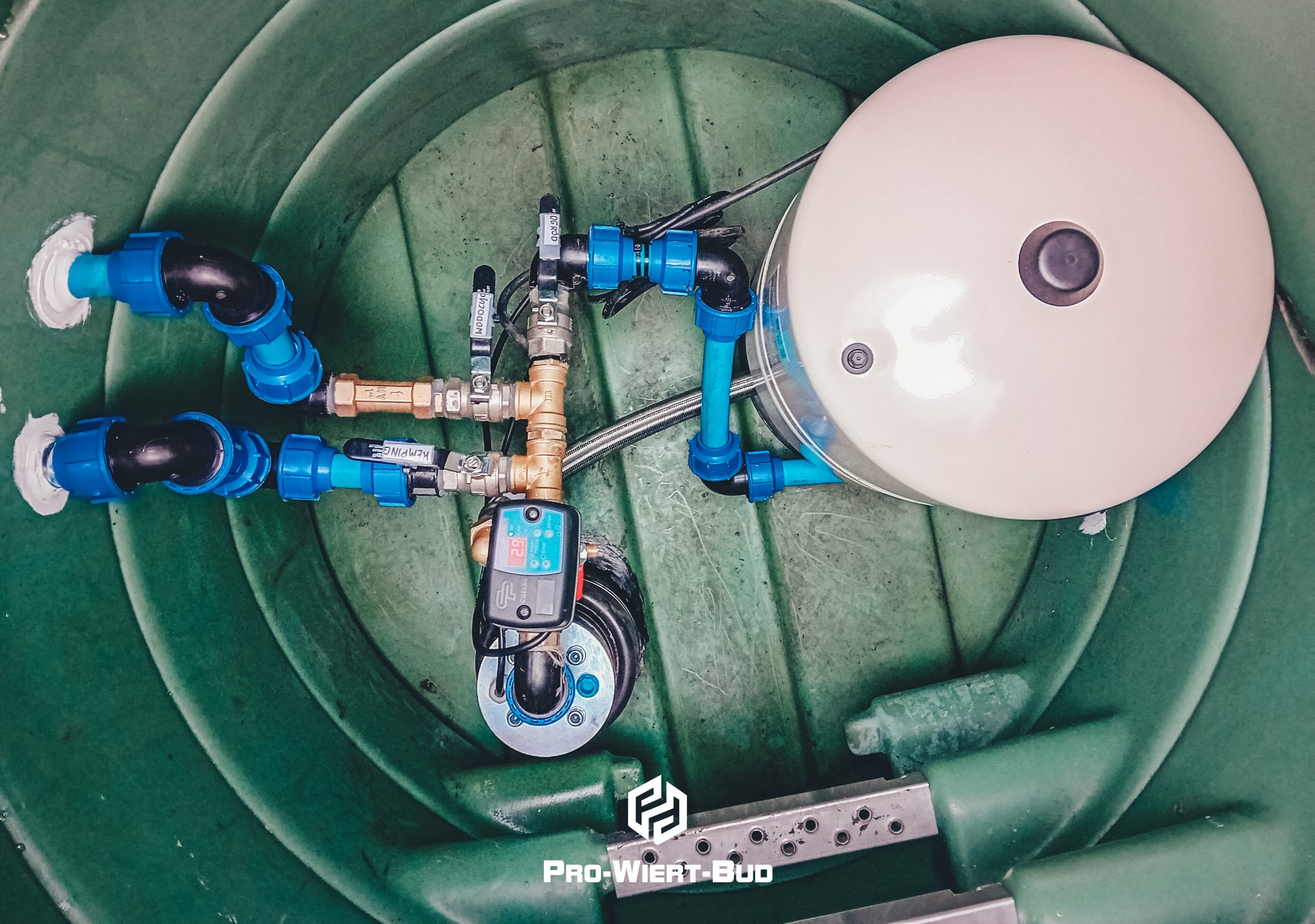 Wnętrze zielonej studni z zamontowanym osprzętem: zbiornik hydroforowy, automat hydroforowy z wyświetlaczem, zawory i niebieskie rury, widoczne logo firmy Pro-Wiert-Bud.