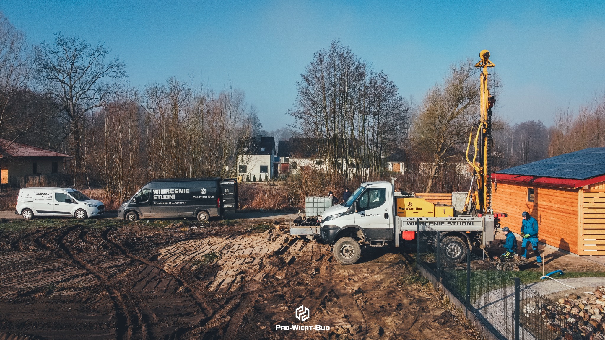 Wiertnica na podwoziu samochodu ciężarowego w trakcie pracy, dwóch pracowników w kombinezonach roboczych, w tle dwa samochody dostawcze z logo firmy, teren budowy z błotnistą ziemią, w oddali...