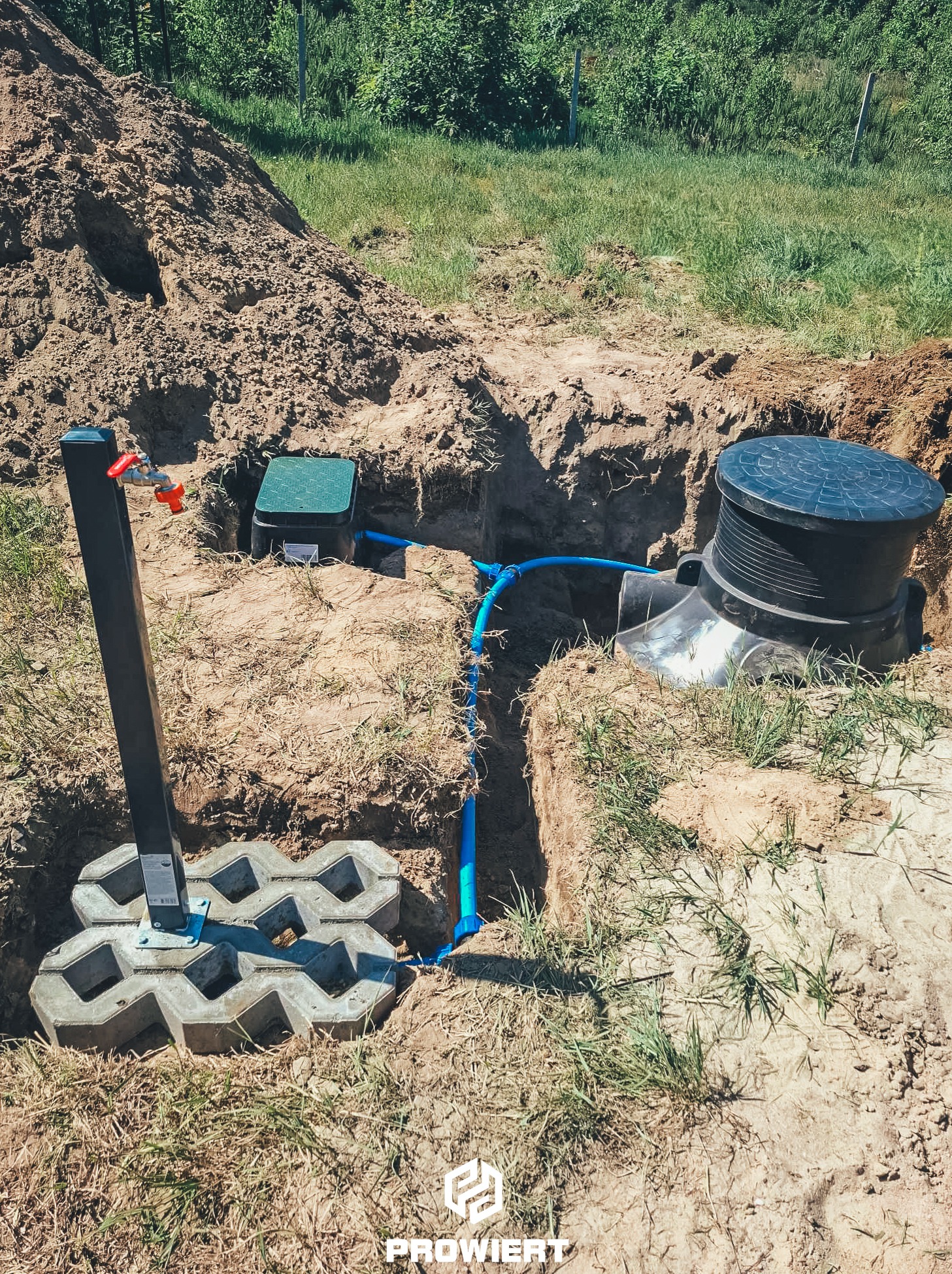 Instalacja studni głębinowej: czarna skrzynka, niebieska rura, kran z czerwoną rączką, obudowa z pokrywą, kostka brukowa, wykop w ziemi, trawa.
