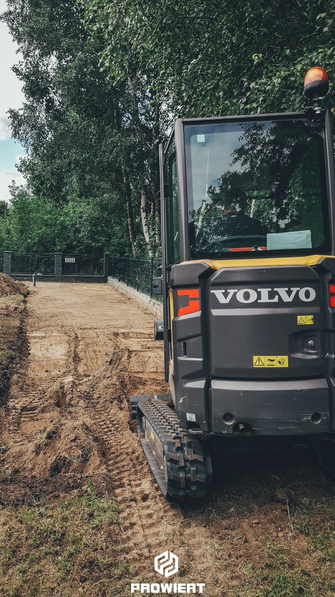 Żółto-czarna koparka Volvo na placu budowy, przygotowującym teren pod studnię. Widoczne ślady opon i świeżo przekopana ziemia. W tle ogrodzenie i zieleń.