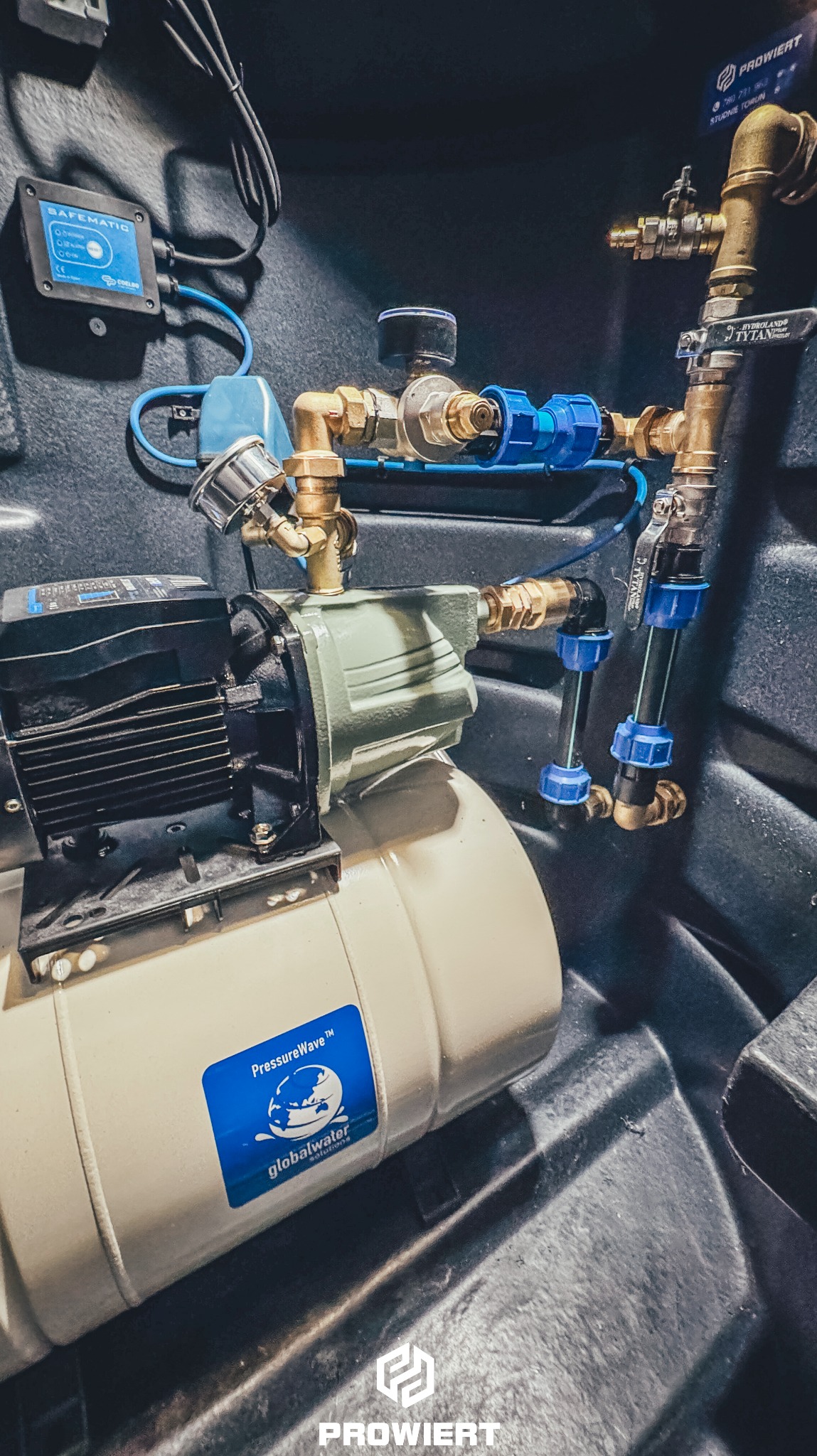 Kompletna instalacja pompy głębinowej w studni z osprzętem: zbiornik hydroforowy, automat SAFEMATIC, zawory i rury. Widok z góry, oświetlenie techniczne.