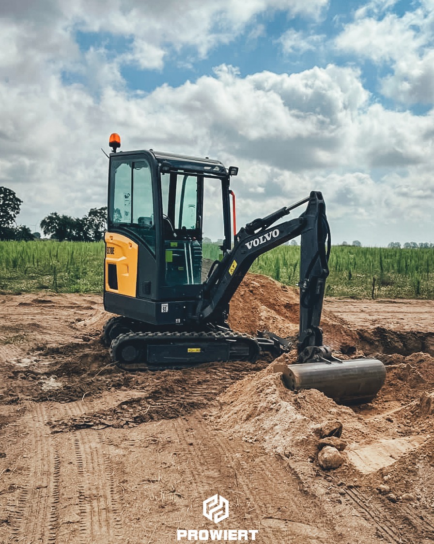 Żółto-czarna minikoparka Volvo na placu budowy, z widocznymi śladami opon w ziemi i stertą piasku, na tle zielonego pola i błękitnego nieba z chmurami.