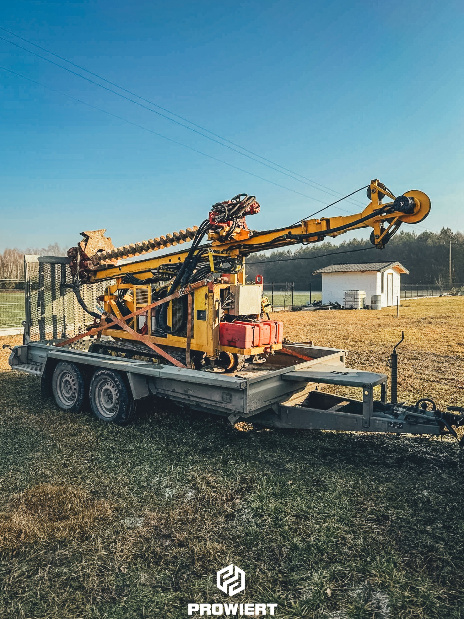 Żółta wiertnica na przyczepie w terenie trawiastym, z białym budynkiem w tle, pod błękitnym niebem. Widok z boku, podkreślający detale maszyny.