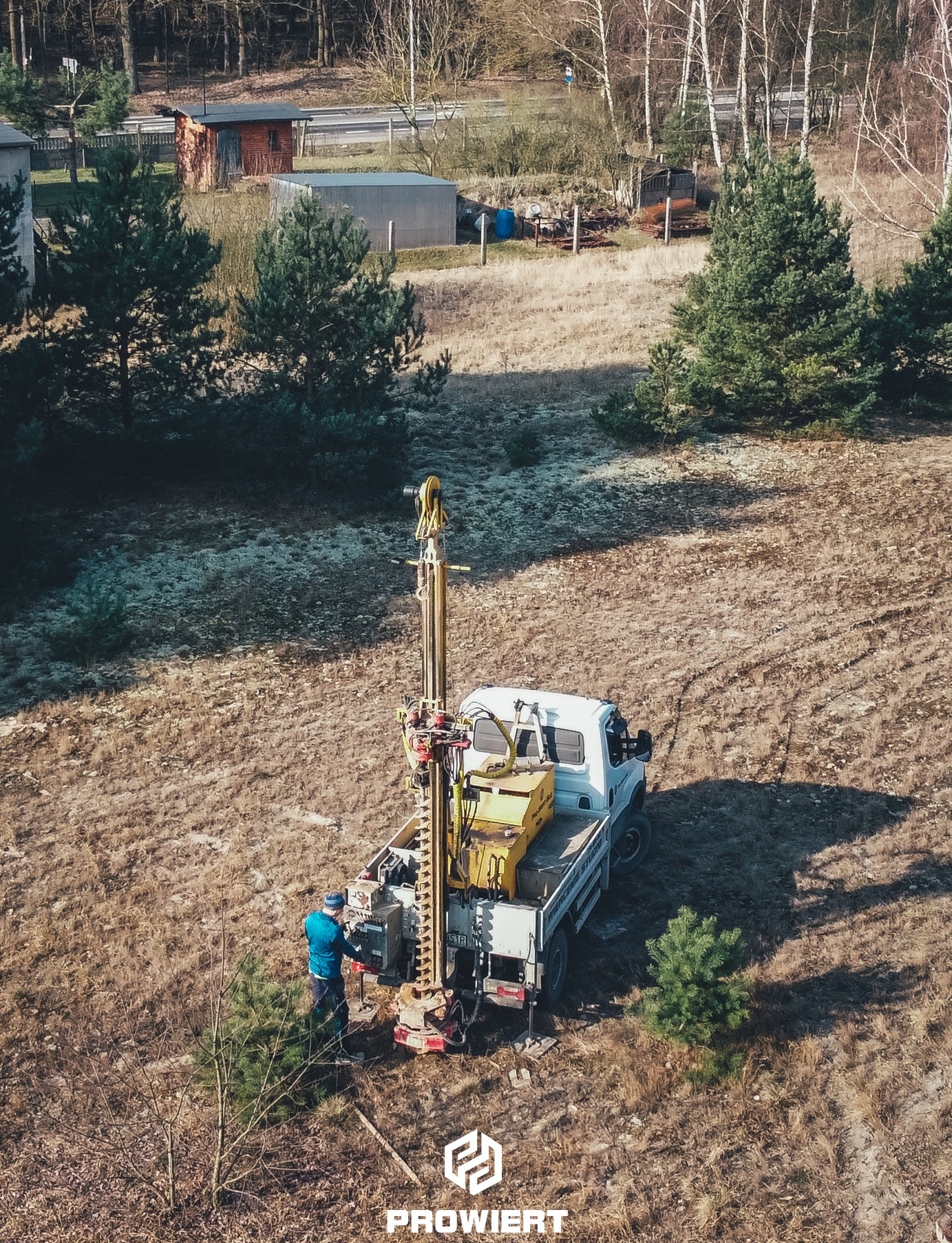 Wiertnica zamontowana na samochodzie ciężarowym podczas pracy na zewnątrz, widok z góry, operator w niebieskiej kurtce, w tle zabudowania i drzewa.