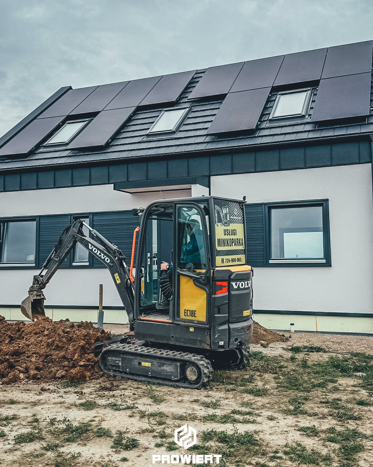 Kompaktowa koparka Volvo EC18E w trakcie prac ziemnych na działce z nowoczesnym domem z panelami słonecznymi na dachu. Widoczny operator w kabinie.