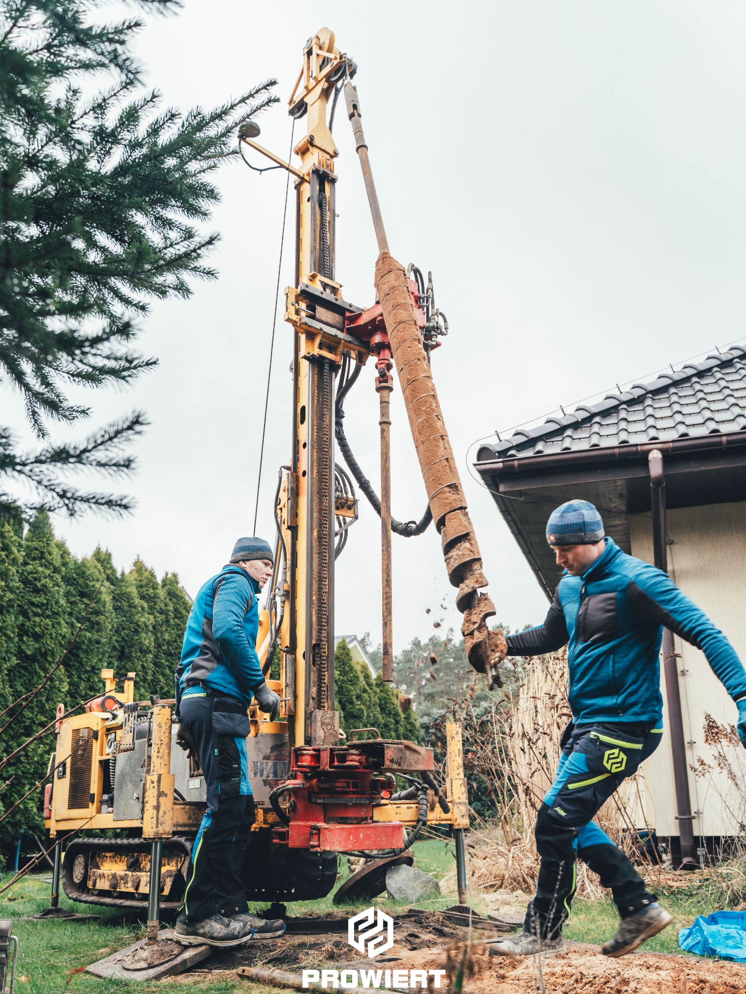 Żółta wiertnica w akcji, dwóch pracowników w niebieskich kurtkach. Wiertło z ziemią uniesione, dom w tle. Praca w toku, dynamiczne ujęcie.