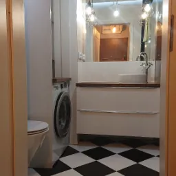 Nowoczesna łazienka z czarno-białą szachownicą na podłodze, białą umywalką nablatową, pralką w zabudowie i wiszącymi lampami w metalowych kloszach.