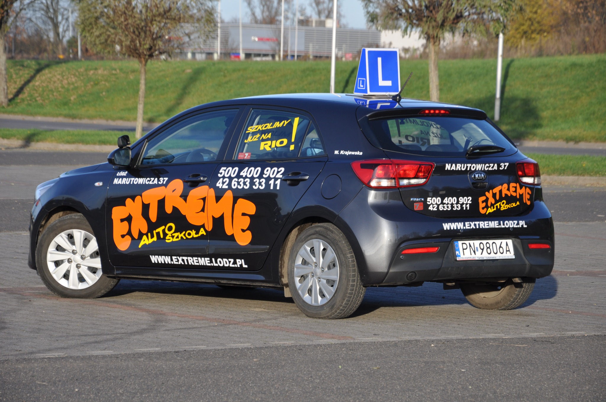 Czarny samochód Kia Rio z oznaczeniami szkoły nauki jazdy Extreme Auto Szkoła, tabliczka 'L' na dachu, zaparkowany na parkingu, w tle trawnik i budynek.