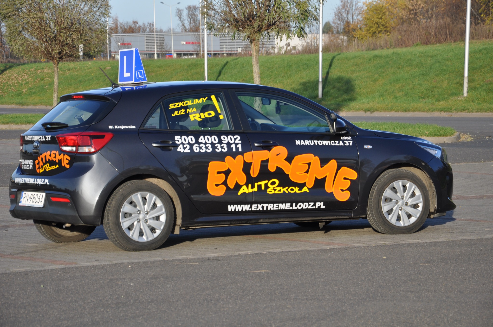 Ciemny samochód z naklejkami 'EXTREME AUTO SZKOŁA' i numerami telefonów, z niebieską literą 'L' na dachu, zaparkowany na parkingu.