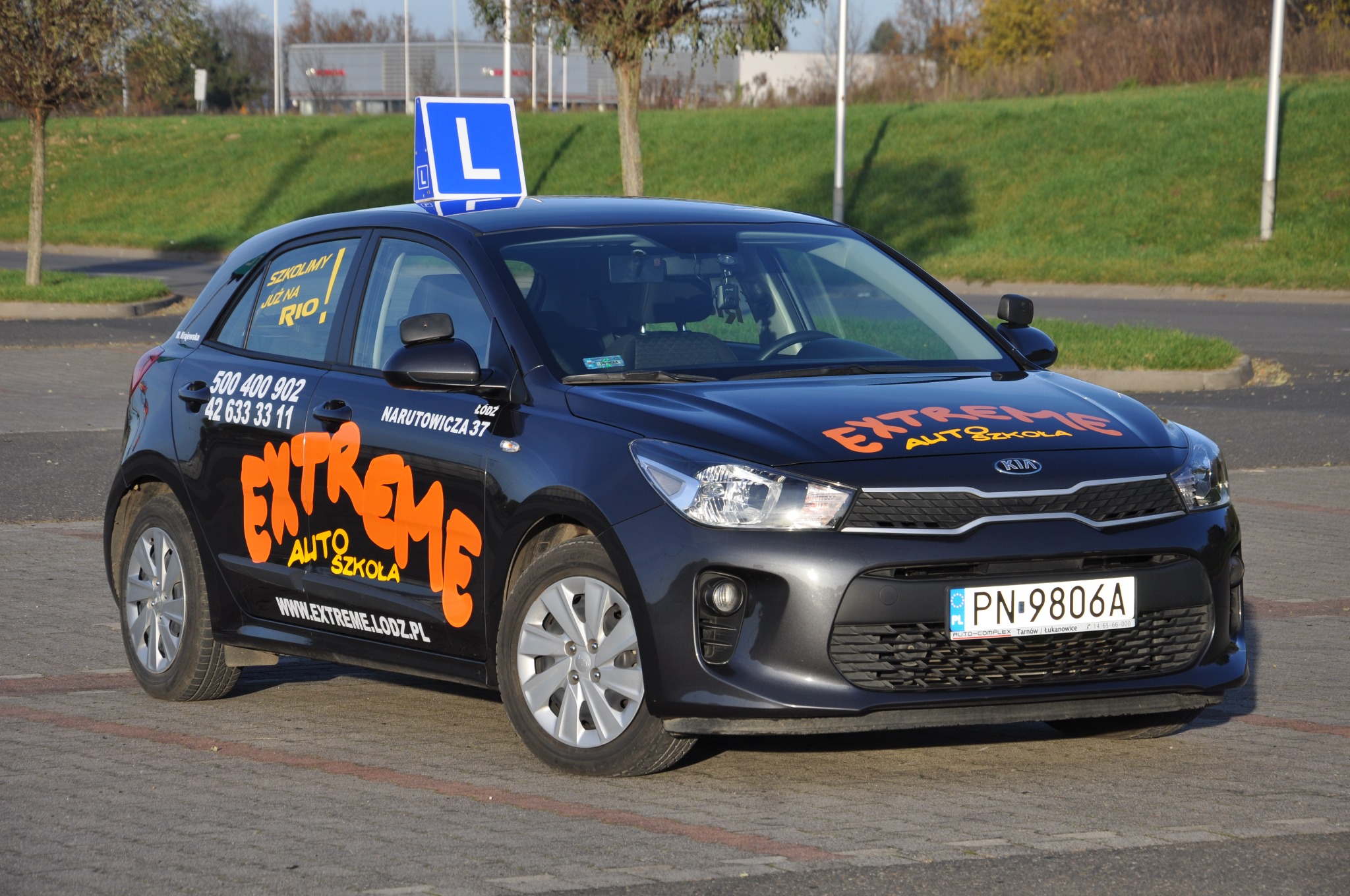 Ciemny samochód Kia Rio z oznaczeniem 'L' na dachu i pomarańczowym napisem 'EXTREME AUTO SZKOŁA' na karoserii, zaparkowany na placu manewrowym szkoły jazdy.