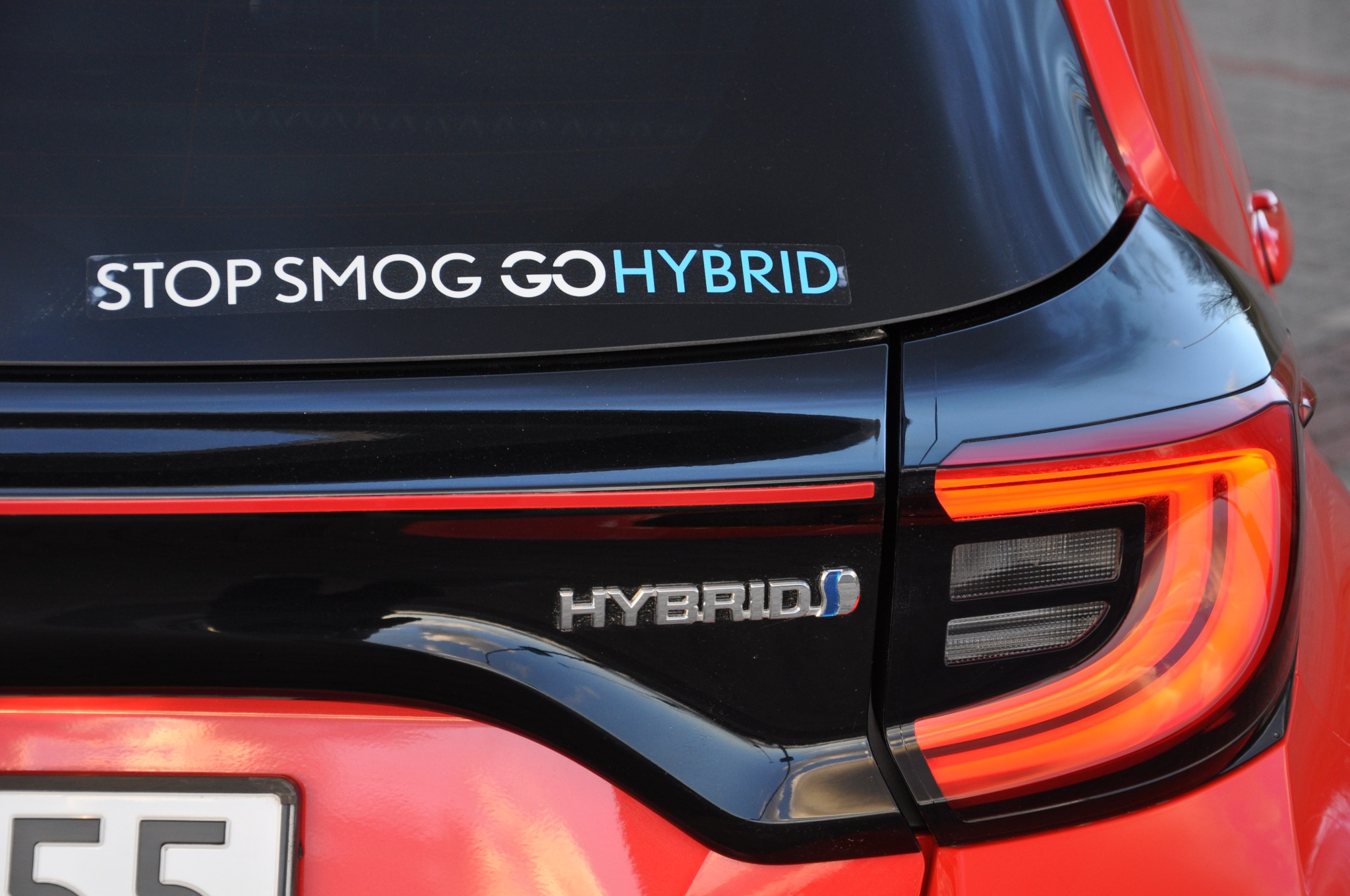 Tył czerwono-czarnego samochodu hybrydowego z naklejką 'STOP SMOG GO HYBRID' na tylnej szybie i widocznym światłem stopu.