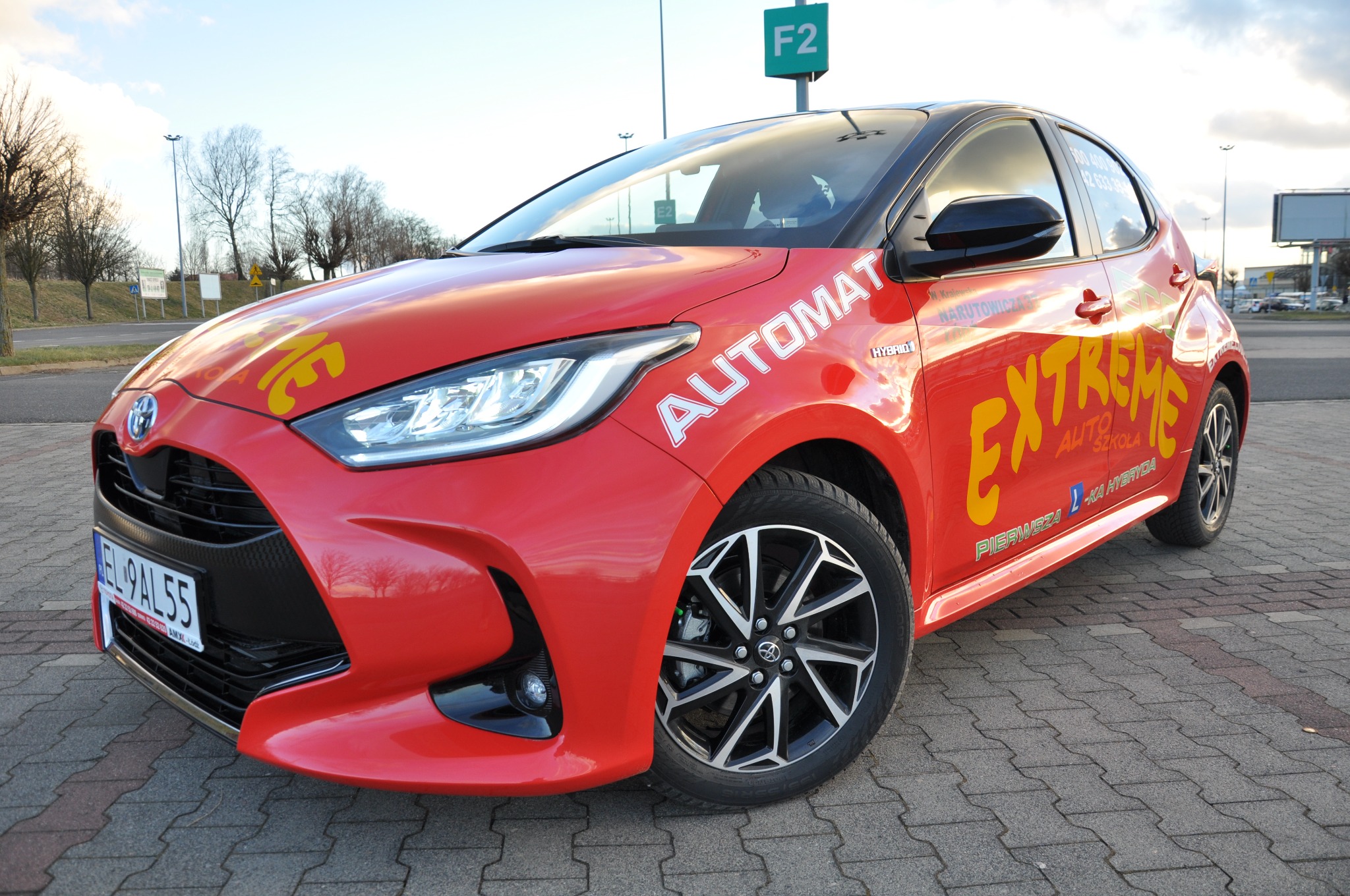 Czerwony samochód Toyota Yaris z oznaczeniami szkoły jazdy Extreme Auto Szkoła, naklejka 'Automat' na przednich drzwiach, zaparkowany na brukowanym parkingu w pochmurny dzień.