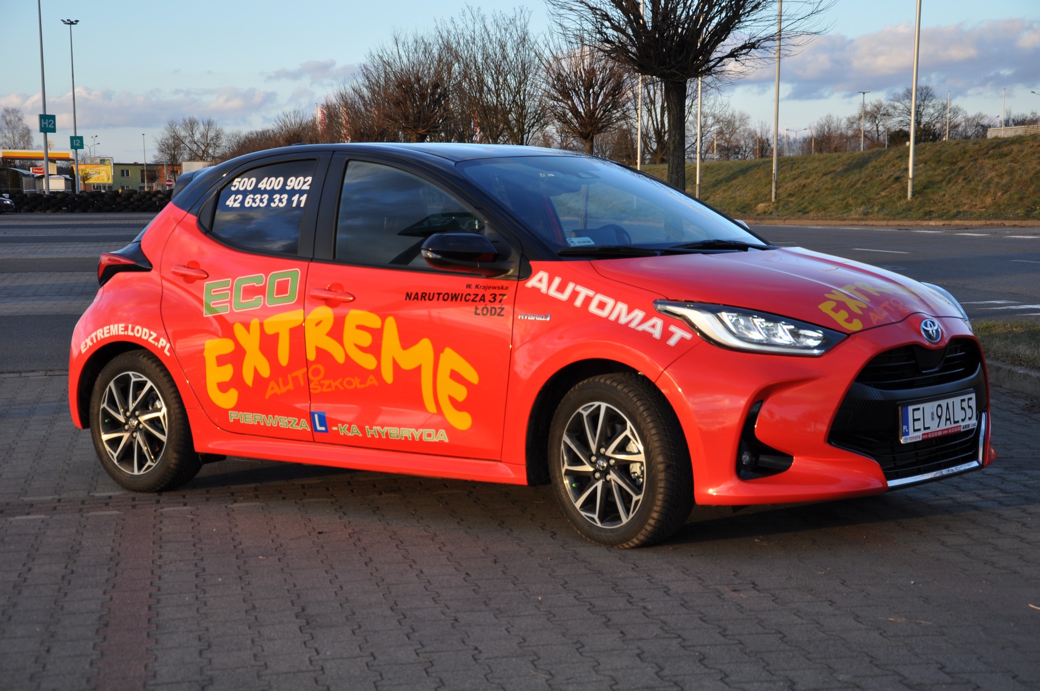 Czerwony samochód z napisem 'Extreme Auto Szkoła' i numerami kontaktowymi, zaparkowany na brukowanej nawierzchni, widok z boku.