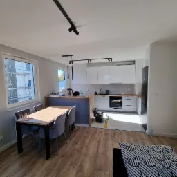 Jasne wnętrze mieszkania po remoncie, widok na kuchnię z białymi szafkami i drewnianym blatem, st&oacute;ł z krzesłami w części jadalnej, podłoga z paneli imitujących drewno, okno z widokiem na rusztowania.