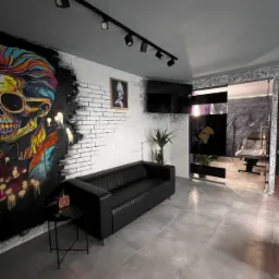 Wnętrze studia tatuażu: czarna skórzana sofa na szarych płytkach, ściana z białej cegły z kolorowym graffiti czaszki, oświetlenie punktowe, czarny stolik boczny z flakonem, widok na stanowisko...