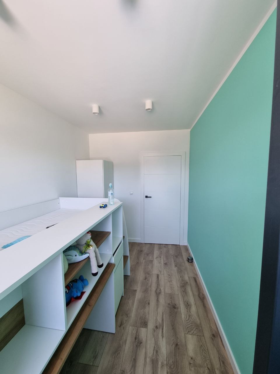 Pokój dziecięcy po remoncie: białe meble z półkami na zabawki, panele podłogowe w odcieniach brązu i turkusowa ściana, oświetlenie punktowe.