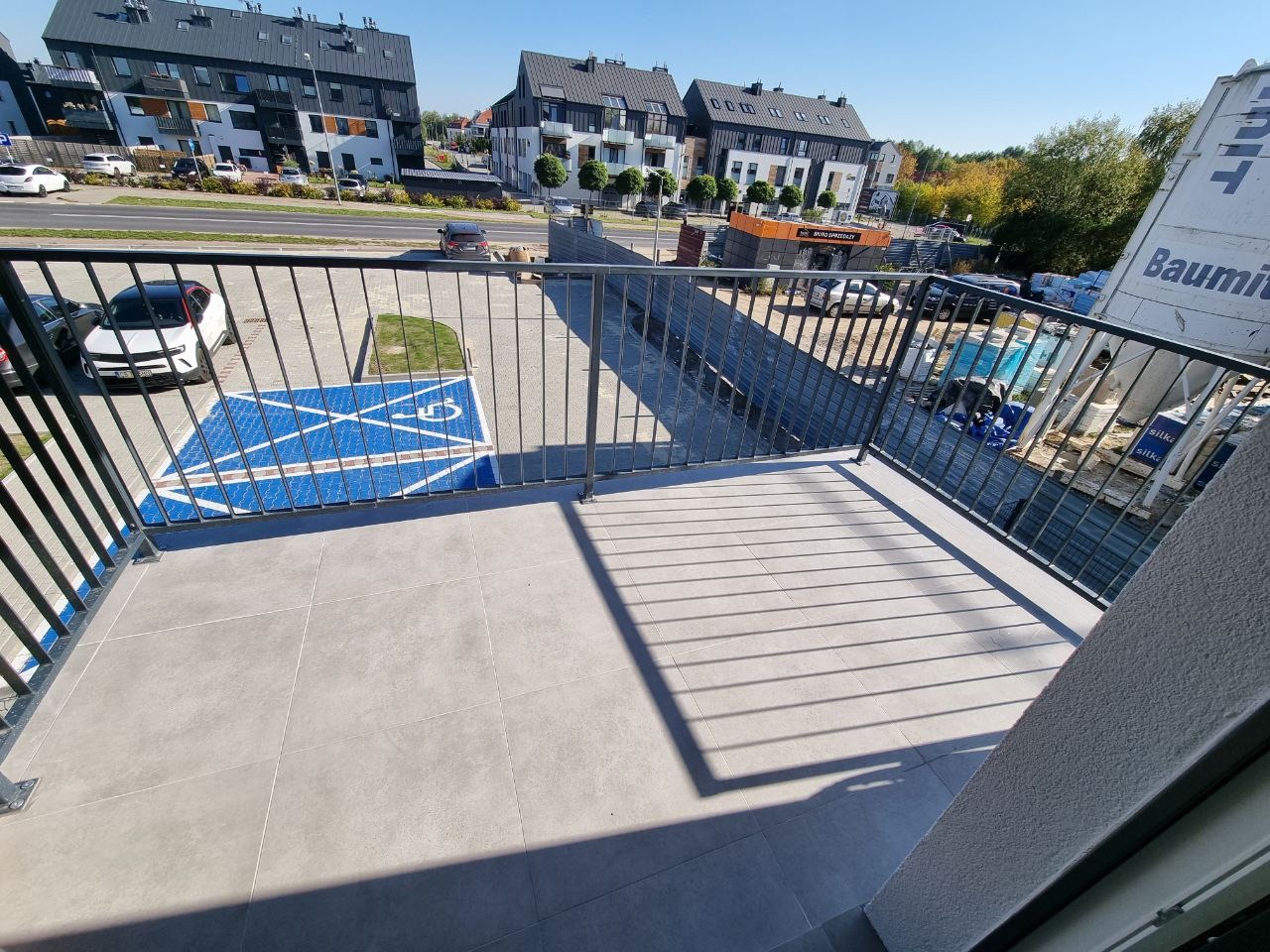 Szare płytki na balkonie w nowym budownictwie, widok na parking z miejscem dla niepełnosprawnych i nowoczesne budynki mieszkalne w tle.