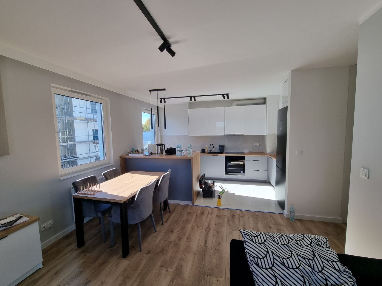 Jasne wnętrze mieszkania po remoncie, widok na kuchnię z białymi szafkami i drewnianym blatem, stół z krzesłami w części jadalnej, podłoga z paneli imitujących drewno, okno z widokiem na rusztowania.