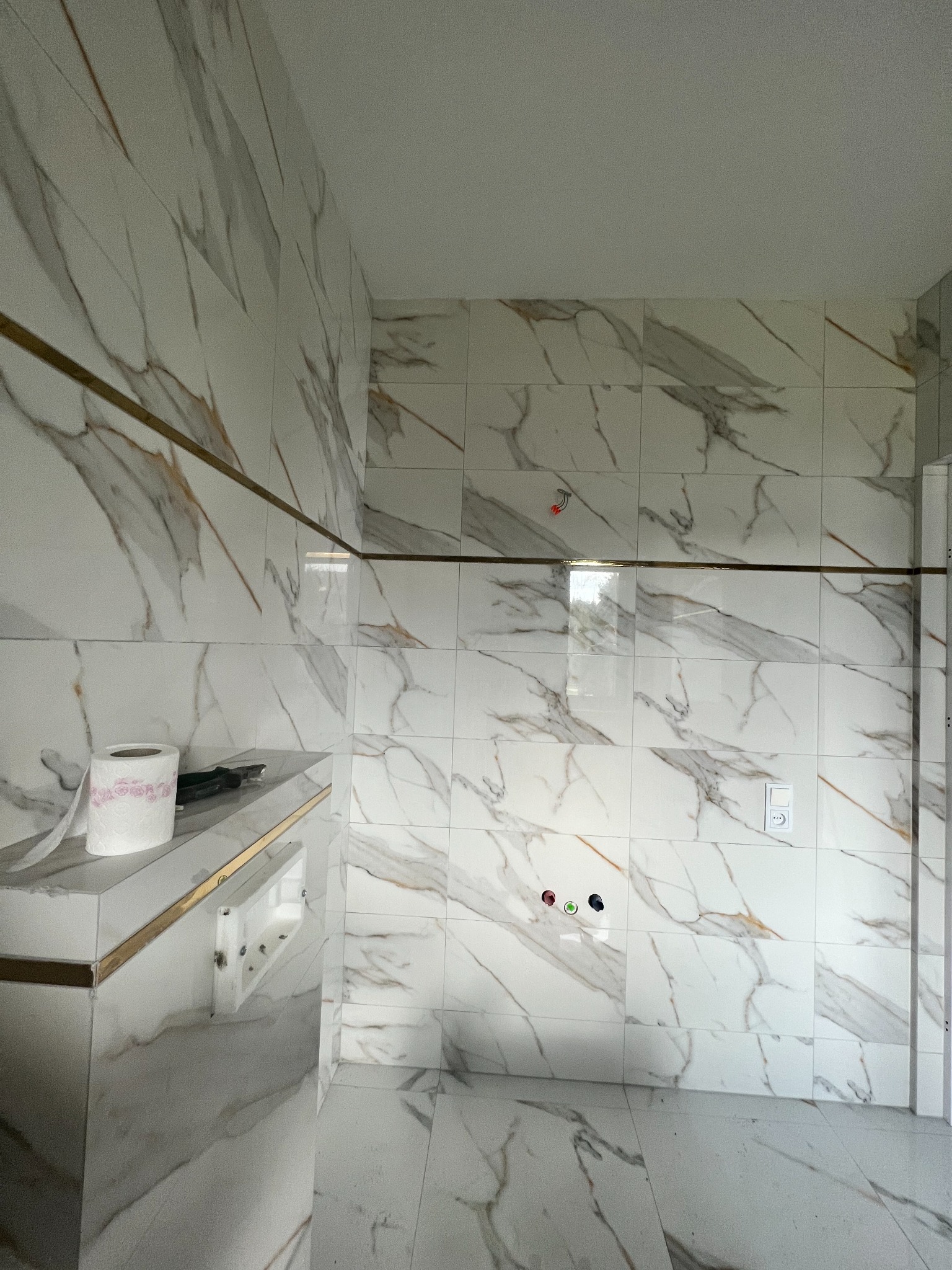 Białe płytki imitujące marmur z złotymi akcentami na ścianach i podłodze w łazience, widoczne przygotowane przyłącza wodne i elektryczne, rolka papieru toaletowego na blacie.
