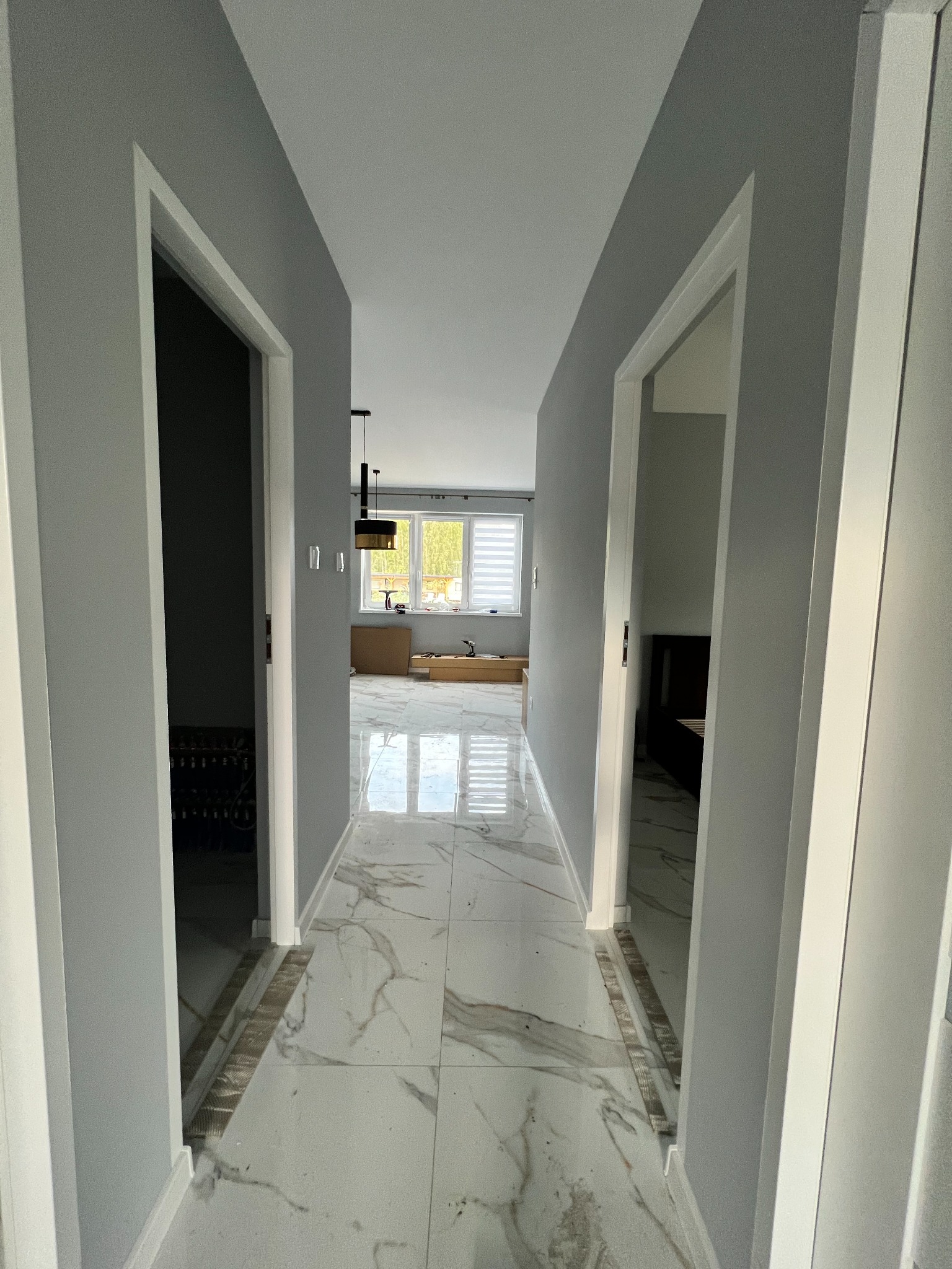 Korytarz w odcieniach szarości z białymi drzwiami i marmurową podłogą, widoczne przygotowania do prac w dalszej części pomieszczenia, w tym narzędzia i materiały budowlane.