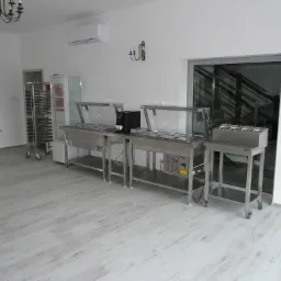 Wyposażenie kuchni remizy OSP: stalowe stoły z podgrzewaczami, chłodziarka i regał na kółkach w jasnym pomieszczeniu z klimatyzacją i schodami w tle.