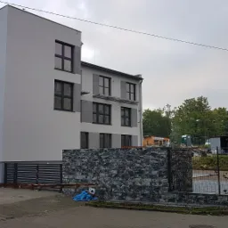 Nowoczesny budynek mieszkalny z szarą elewacją i czarnymi ramami okiennymi, ogrodzony murem z szarej kostki. Na drugim planie widoczny starszy blok i teren budowy.
