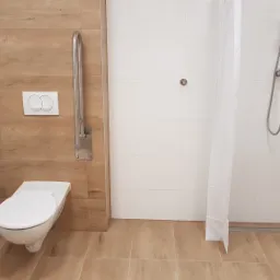 Wykończona łazienka z udogodnieniami dla osób z niepełnosprawnościami: poręcze przy toalecie i umywalce, natrysk bez brodzika z siedziskiem, białe i drewnopodobne płytki.