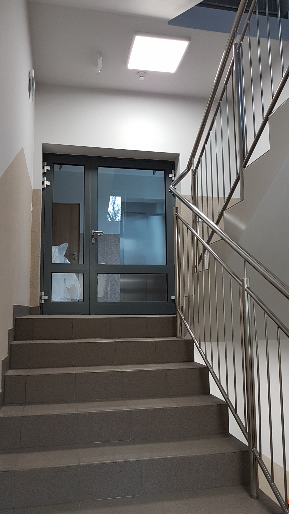 Wewnętrzne schody z szarych płytek prowadzące do przeszklonych, grafitowych drzwi z metalowymi okuciami i poręczą ze stali nierdzewnej.