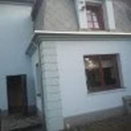 Arkadiusz Matuszak - Odnowiona elewacja budynku w Warszawie z nową, jasną farbą, widoczne okno z ciemną ramą i fragment dachu. Część ściany wykończona dekoracyjną okładziną imitującą kamień.